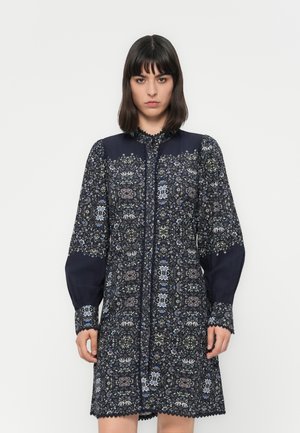 Una donna con capelli scuri indossa un vestito blu navy a maniche lunghe e lunghezza al ginocchio, decorato con intricati motivi floreali e rifiniture in pizzo, su uno sfondo bianco semplice.