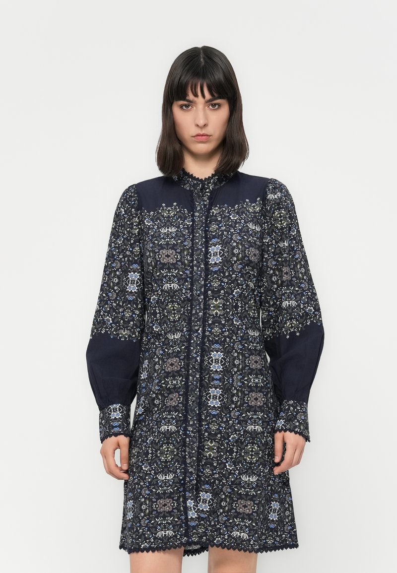 Woman with dark hair wears a long-sleeve, knee-length navy dress with intricate floral patterns and lace trim against a plain white background.