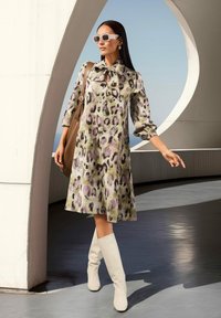Frau, die ein gemustertes Kleid mit einer Schleife, weiße Stiefel, Sonnenbrille trägt und eine braune Tasche hält, steht unter einer geschwungenen architektonischen Struktur.