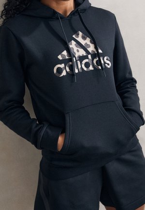 Persoon die een zwarte Adidas hoodie draagt met een grijs luipaardprintlogo en bijpassende zwarte korte broek, handen in de zak van de hoodie.