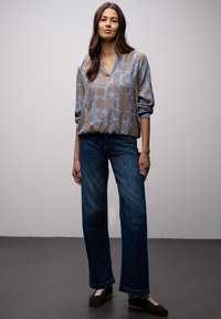 Femme aux longs cheveux bruns portant un chemisier à motifs géométriques bleu et beige, un jean bleu et des baskets foncées, debout sur un sol gris.