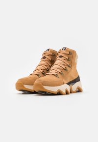 Høye støvler i kaki semsket skinn med beige detaljer, strukturert snøring og en tykk hvit og brun såle. Har en logo på hælen.