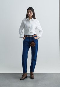 Camicia bianca semi-adattata con dettagli arricciati, abbinata a jeans blu a vita alta, cintura marrone e stivaletti alla caviglia coordinati.