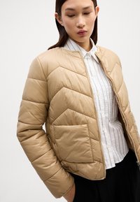 Beige pufferjakke med et chevron-quiltet design, lynlås i front og en lomme, båret over en hvid stribet skjorte. Glat tekstur.