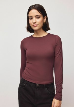 Pika pikkade varrukatega marooni värvi topp, mis on valmistatud venivast kangast. Omab ümmargust kaelust ja keha järgivat siluetti, mida on kombineeritud mustade pükstega.