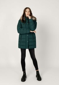 Giacca imbottita verde scuro con cappuccio foderato in pelliccia, bottoni a pressione e due tasche con zip. Indossata sopra jeans skinny neri e stivali neri voluminosi.