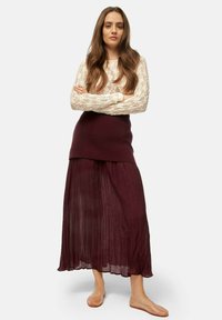 Burgundy pleated maxi skirt paired with a cream lace long-sleeve top, complemented by nude flat shoes. Textured fabric, layered design.