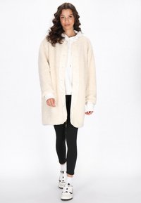 Donna in giacca di pile beige chiaro con bottoni, felpa bianca con cappuccio, leggings neri e sneakers bianche che cammina su uno sfondo bianco uniforme.