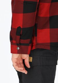 Rood en zwart geruit button-up jack met een omgeslagen mouw, voorzien van een metalen drukknoop. Zwarte broek met een beige merklabel op de zak.