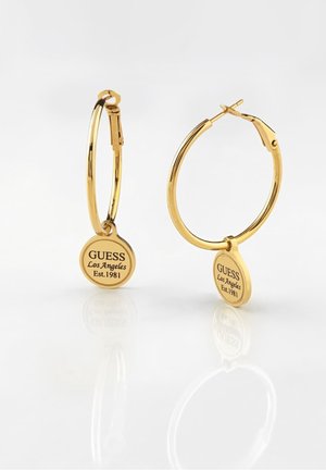 Goldene Creolen mit runden Anhängern, graviert mit "GUESS Los Angeles Est. 1981" auf einer reflektierenden weißen Oberfläche.
