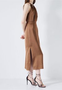 Robe brune sans manches avec un décolleté tordu, présentant une fente latérale et une texture lisse. Associée à des sandales à talons hauts noires à lanières.