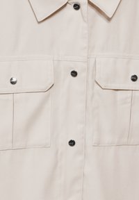 Vaalean beige nappipaita, joka on valmistettu tasaisesta kankaasta. Paidassa on kaksi rintataskua ja hopeanväriset napit. Klassinen kaulus ja tekstiilistruktuuri.