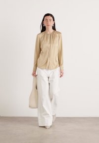 Blusa di seta color tan con dettagli arricciati, abbinata a pantaloni bianchi a gamba larga e una borsa a tracolla beige. La modella indossa calzature neutre.