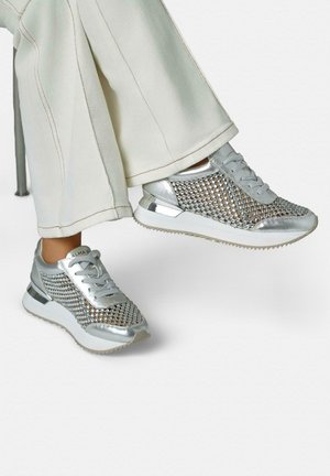 Personne portant un pantalon blanc et des baskets en maille argentée ornées de strass, assise sur une chaise grise.