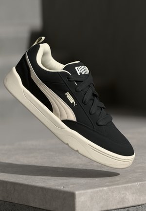 Zwarte en crèmekleurige sneaker met een gladde textuur, een wit Puma-logo en bijpassende veters; heeft een gewatteerde kraag en een rubberen zool.