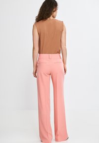 Kobieta z kręconymi włosami, ubrana w bezrękawnik w kolorze brązowym oraz różowe spodnie z wysokim stanem i szerokimi nogawkami, stojąca na tle białej ściany.