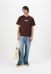 Young man wearing a brown graphic T-shirt, loose blue jeans, and brown sneakers, holding a beige tote bag against a white background.