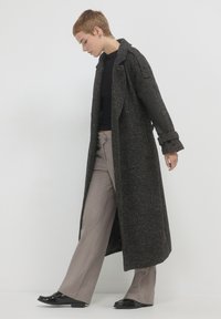 Lungo cappotto nero testurizzato con ampi revers, taglio oversize e maniche arrotolate, indossato su una maglia nera e pantaloni in corduroy beige.