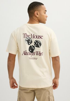 Hombre con camisa beige con gráfico de dados y texto "La casa siempre gana" y texto más pequeño "Juega cada partida como si fuera la última."