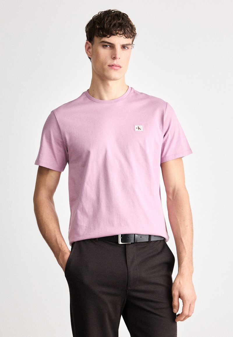 Rosa bomulls t-skjorte med korte ermer og rund hals; har et lite logopatch på brystet. Brukt med mørke, tettsittende bukser og et svart belte.