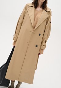 Beige trench coat with a double-breasted front, black buttons, wide lapels, and oversized sleeves, paired with a beige sweater and black tote bag.