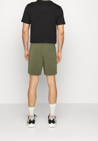 T-shirt noir à manches courtes accompagné d'un short vert olive, avec une poche arrière, des chaussettes blanches et des chaussures de sport noires et rouges.