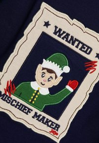 Der gestickte Patch zeigt einen ungezogenen Elfen in einem grünen Outfit mit weißen Akzenten und einem roten Handschuh, umrahmt von einem Design eines gesuchten Posters.