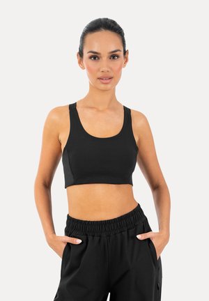 Kvinde med mørkt hår trukket tilbage iført sort sports-bh og sorte atletikbukser med elastisk talje, stående med hænderne i lommerne mod en ensfarvet baggrund.