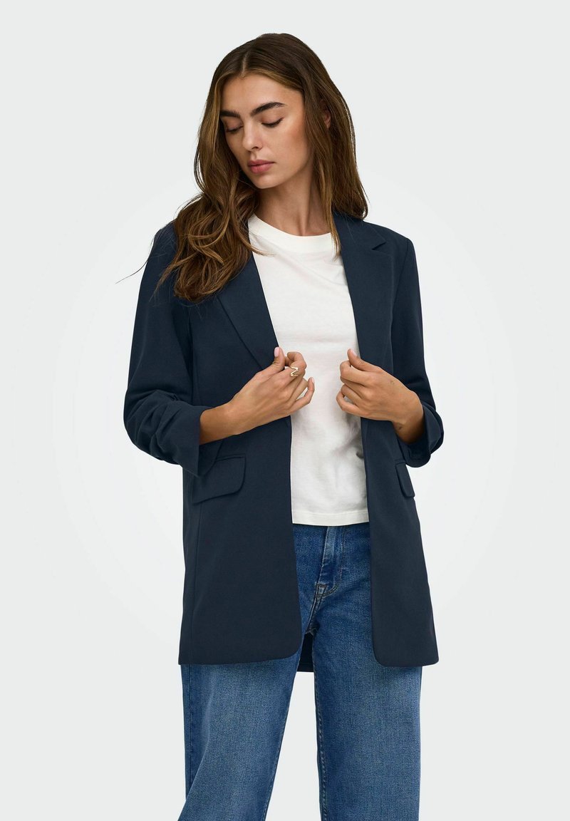 Femme aux longs cheveux bruns portant un blazer bleu marine par-dessus une chemise blanche et un jean bleu, ajustant le blazer avec ses deux mains.