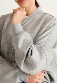 Šedá mikina s kulatým výstřihem a žebrovanými manžetami. Je vyrobena z měkké, texturované tkaniny a obsahuje jemné švy na přední straně.