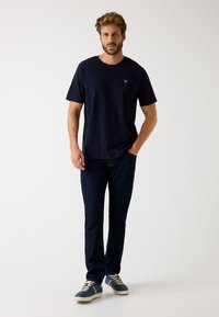 T-shirt in cotone blu navy con scollatura a girocollo e un piccolo logo sul lato sinistro del petto, abbinata a jeans blu scuro e sneakers blu.