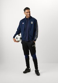 Veste de survêtement marine avec zip, logo sur la poitrine et manches à motifs. Pantalons de sport noirs avec des accents bleus et chaussures de football noires. Tient un ballon de football.