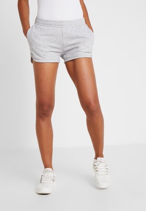 Persoon die lichtgrijze shorts en witte sneakers draagt, staat tegen een eenvoudige witte achtergrond met handen in de zakken.