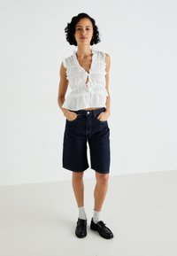 White, ruffled sleeveless top with a tie front, paired with dark denim shorts. Model wears black shoes and gray socks. Simple background.