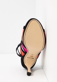 Semelle d'une sandale à talons hauts pour femme avec des sangles noires et des touches orange et rose, taille 37, positionnée contre un mur blanc d'angle.