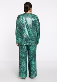 Vrouw met donker haar in een lage knot, gekleed in een glanzende groene en zwarte dierenprint top en brede broek, staand met haar rug naar de camera op een witte achtergrond.