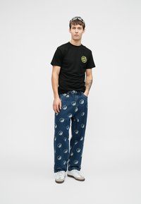 Μαύρη βαμβακερή μπλούζα με κίτρινο κυκλικό λογότυπο, σε συνδυασμό με μπλε τζιν παντελόνι με εκτύπωση yin-yang, φορεμένο με άσπρα αθλητικά παπούτσια.