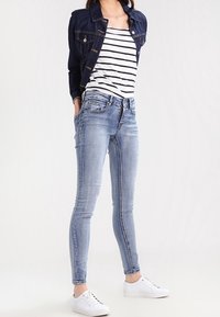 Jeans in denim blu skinny con una leggera scoloritura, abbinati a una canotta a righe bianche e blu navy e a un giubbotto in denim scuro, completati da sneaker bianche.