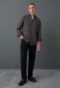 Checkered shirt in dark burgundy with white and green lines, worn over a gray t-shirt; black pants and dark shoes complete the look.