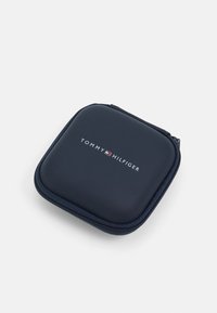 Custodia blu navy con zip realizzata in materiale liscio, con il logo "TOMMY HILFIGER" in bianco e accenti rossi nella parte superiore. Forma quadrata compatta.