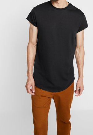 Black cotton t-shirt with short sleeves, round neckline, and a curved hem. Worn with brown pants, highlighting the t-shirt's fit and design.