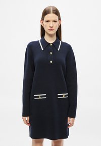 Tengerészkék kötött ruha galléros nyakkivágással, arany gombokkal és két első zsebbel, melyeken fehér csíkos díszítések találhatók. Hosszú ujjú, bő szabású.