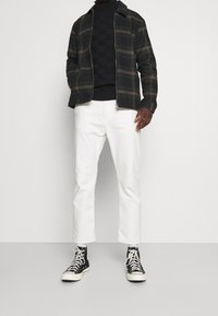 Muž v bílých kalhotách, černozelené kárované bundě, černém kostkovaném svetru a černých vysokých teniskách s bílými tkaničkami.