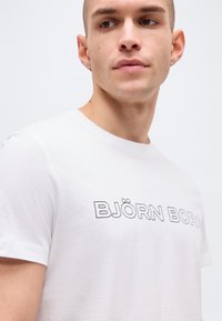 Νεαρός άνδρας με ξυρισμένο κεφάλι που φοράει λευκό μπλουζάκι Björn Borg, κοιτάζοντας ελαφρώς προς το πλάι σε απλό φόντο.