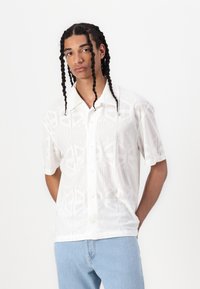 Chemise blanche à manches courtes avec un motif géométrique en relief, dotée d'un col boutonné et d'une coupe décontractée. Portée avec un jean bleu clair.