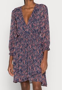 Kleid mit Blumenmuster, tiefem V-Ausschnitt, langen Puffärmeln und gerafftem Taillenzug. Hergestellt aus leichtem Stoff in Marineblau mit pinken Blumen.