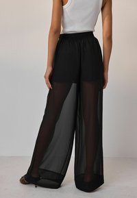 Black sheer wide-leg pants with an elastic waistband, featuring a lightweight fabric that drapes fluidly. Open hem with a smooth texture.