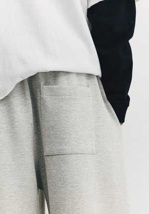 Pantaloni de trening din bumbac, de culoare gri deschis, cu buzunar lateral, având o textură netedă și potrivire lejeră, asortați cu un top alb și negru.