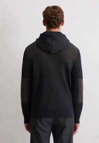 Junger Mann mit kurzem, welligem Haar, der einen schwarzen, strukturierten Kapuzenpullover und dunkle graue Hosen trägt und vor einer schlichten weißen Wand steht.