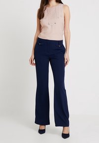 Světle růžový top bez rukávů s korálkovými ozdobami, doplněný tmavě modrými rozšířenými kalhotami a černými lodičkami.